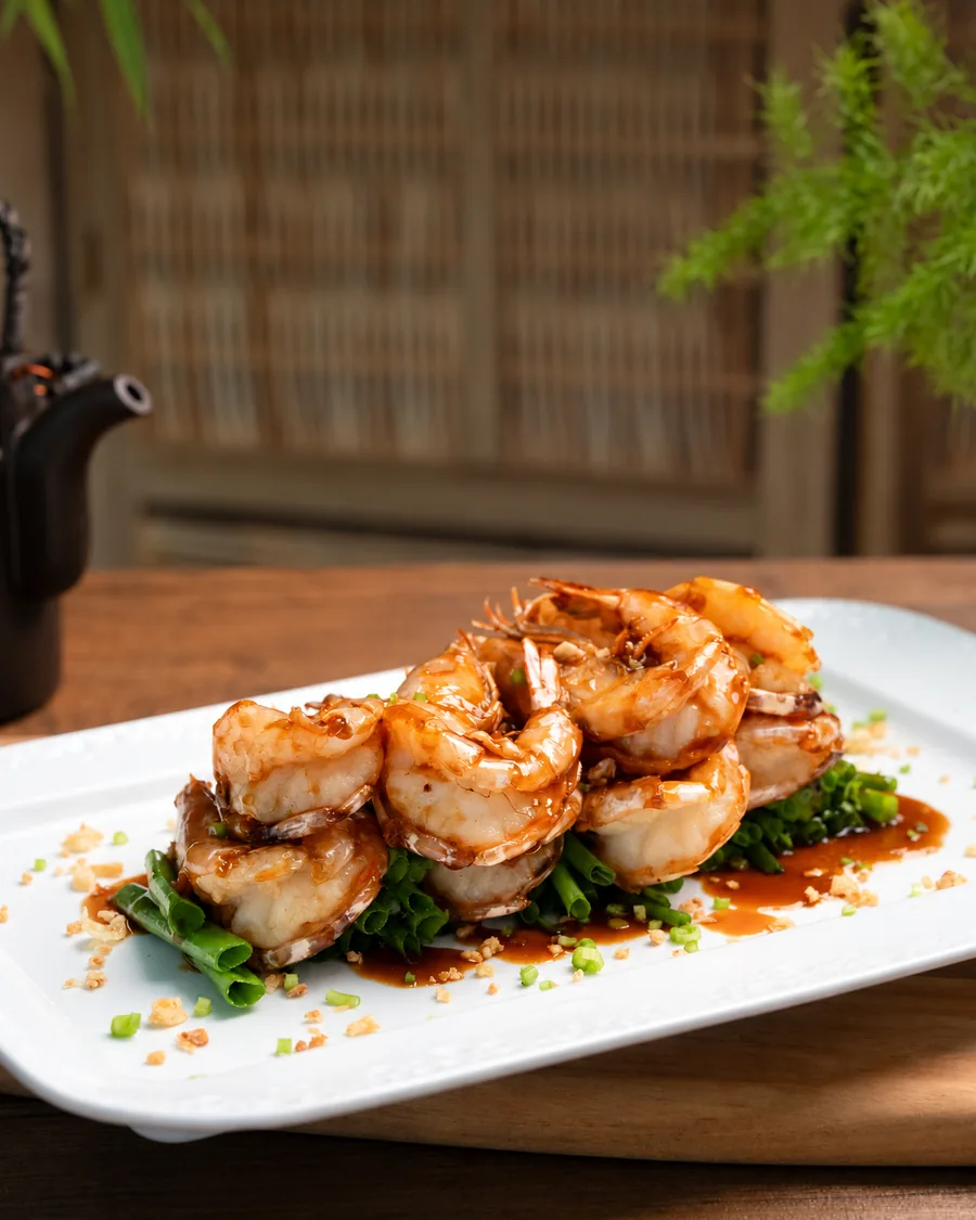 Pan-fried crystal prawns glazed in a glossy oyster sauce on spring onions, served on a white plate